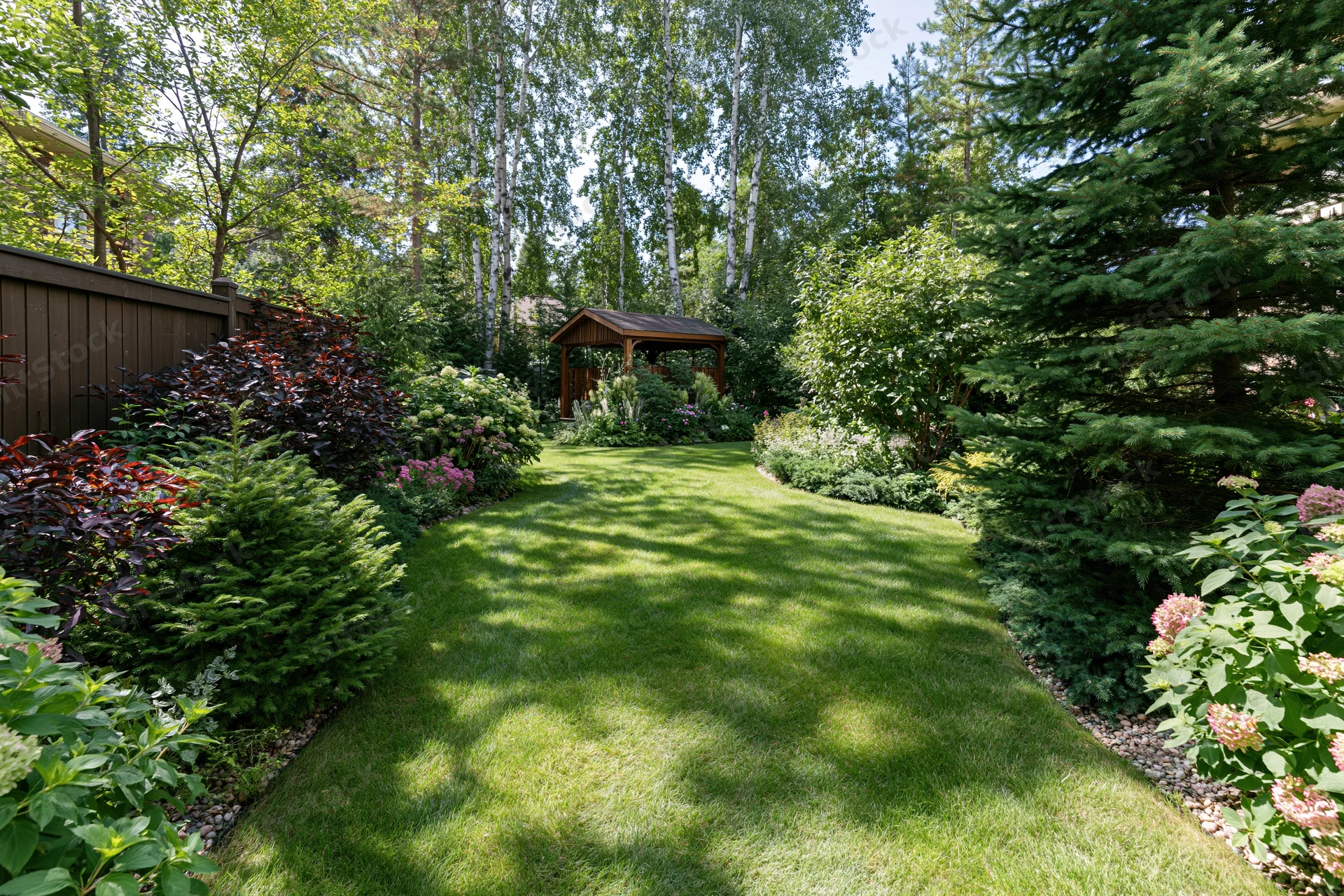 A narrow green lawn path through a lush backyard garden bordered by shrubs and trees leading to a wooden gazebo