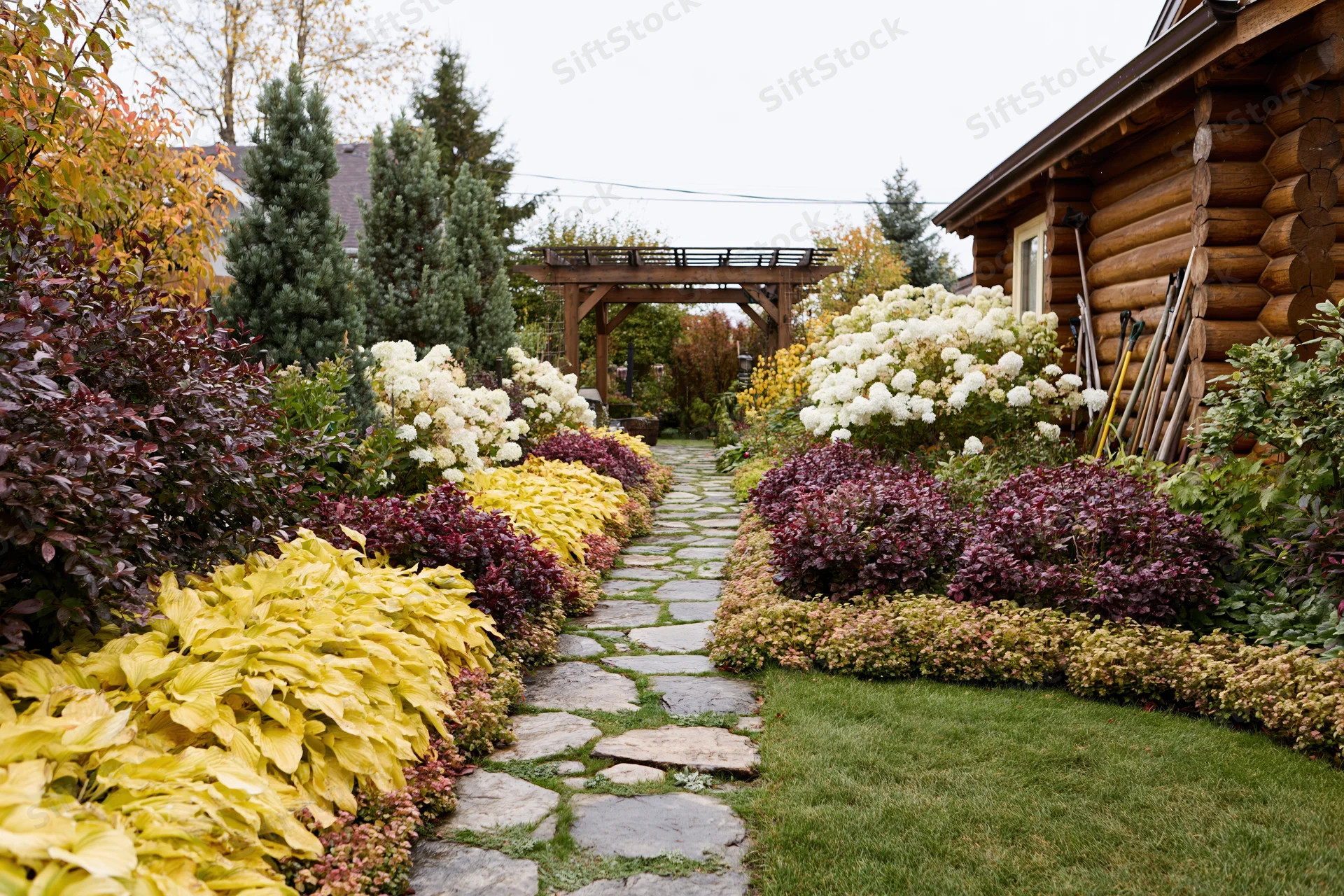 Stone path lined with colorful autumn plants leading through a backyard garden beside a log cabin