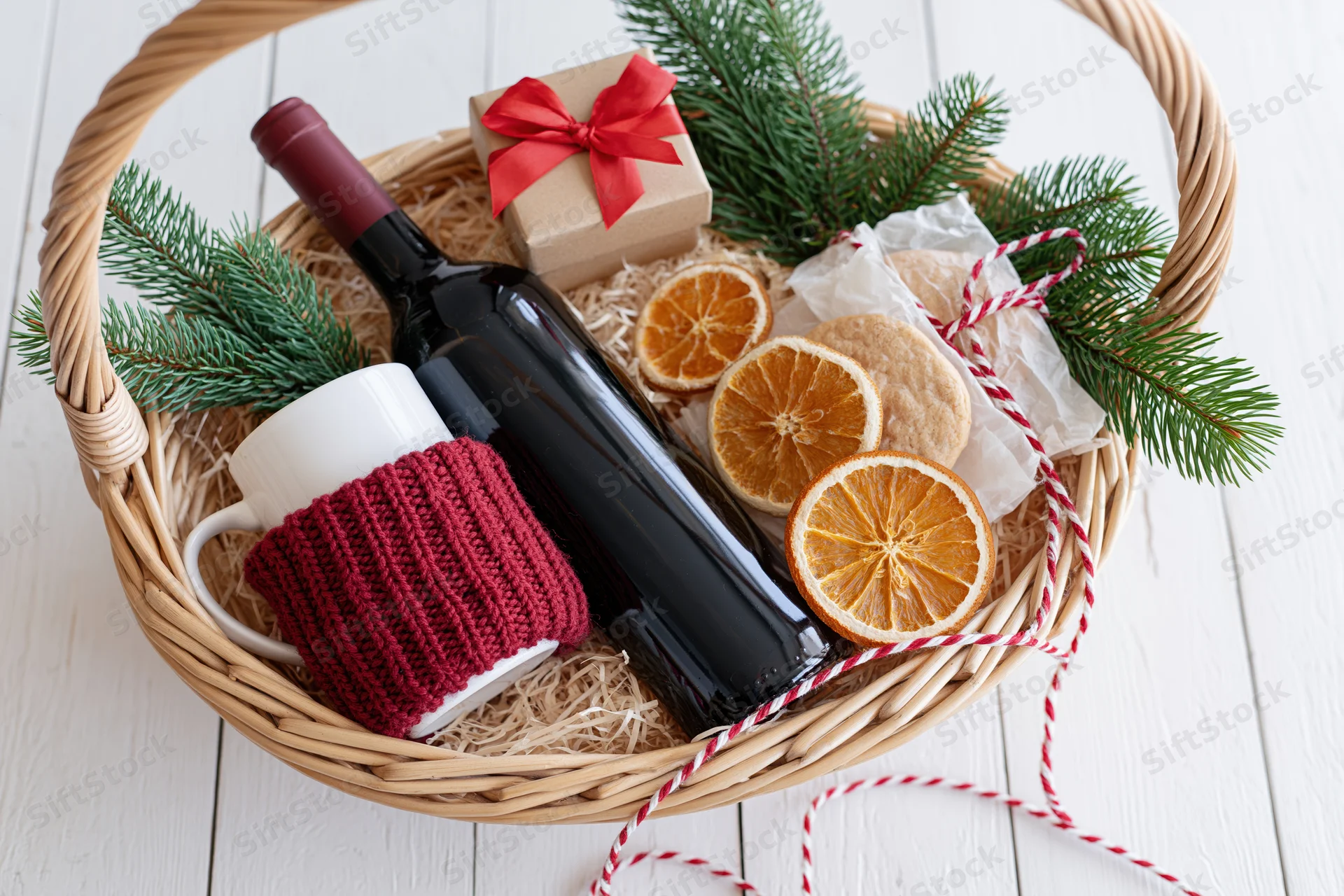 Wicker gift basket filled with red wine bottle, white mug with red knitted cozy, dried orange slices, pine sprigs, small gift box, and red-and-white twine on white wooden surface