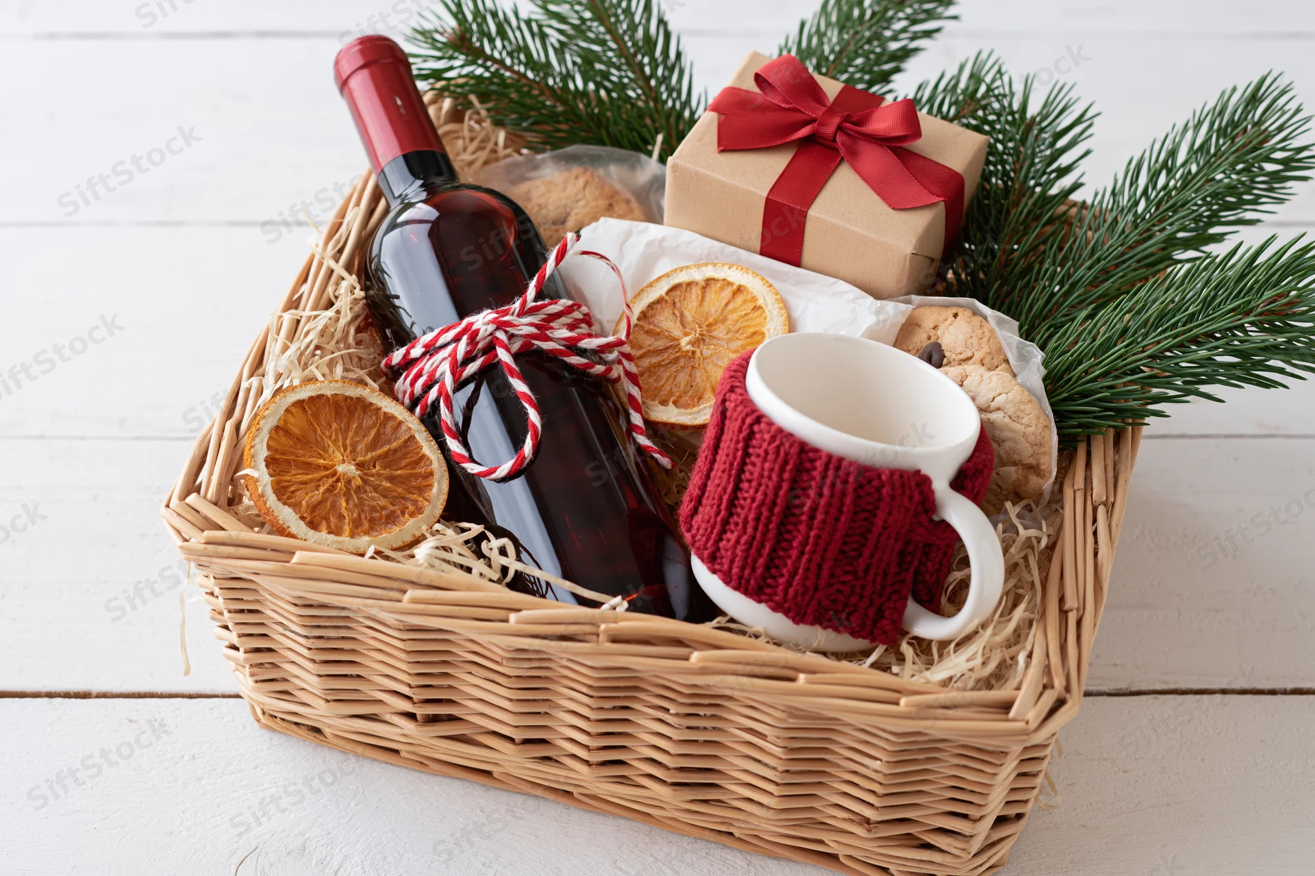 Wicker gift basket filled with red wine bottle, white mug in red knitted cozy, cookies, dried orange slices, and pine branches on white wooden surface