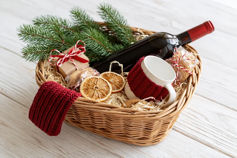 Wicker gift basket filled with wine bottle, white mug, dried oranges, pine sprigs, and wrapped gifts on wooden table