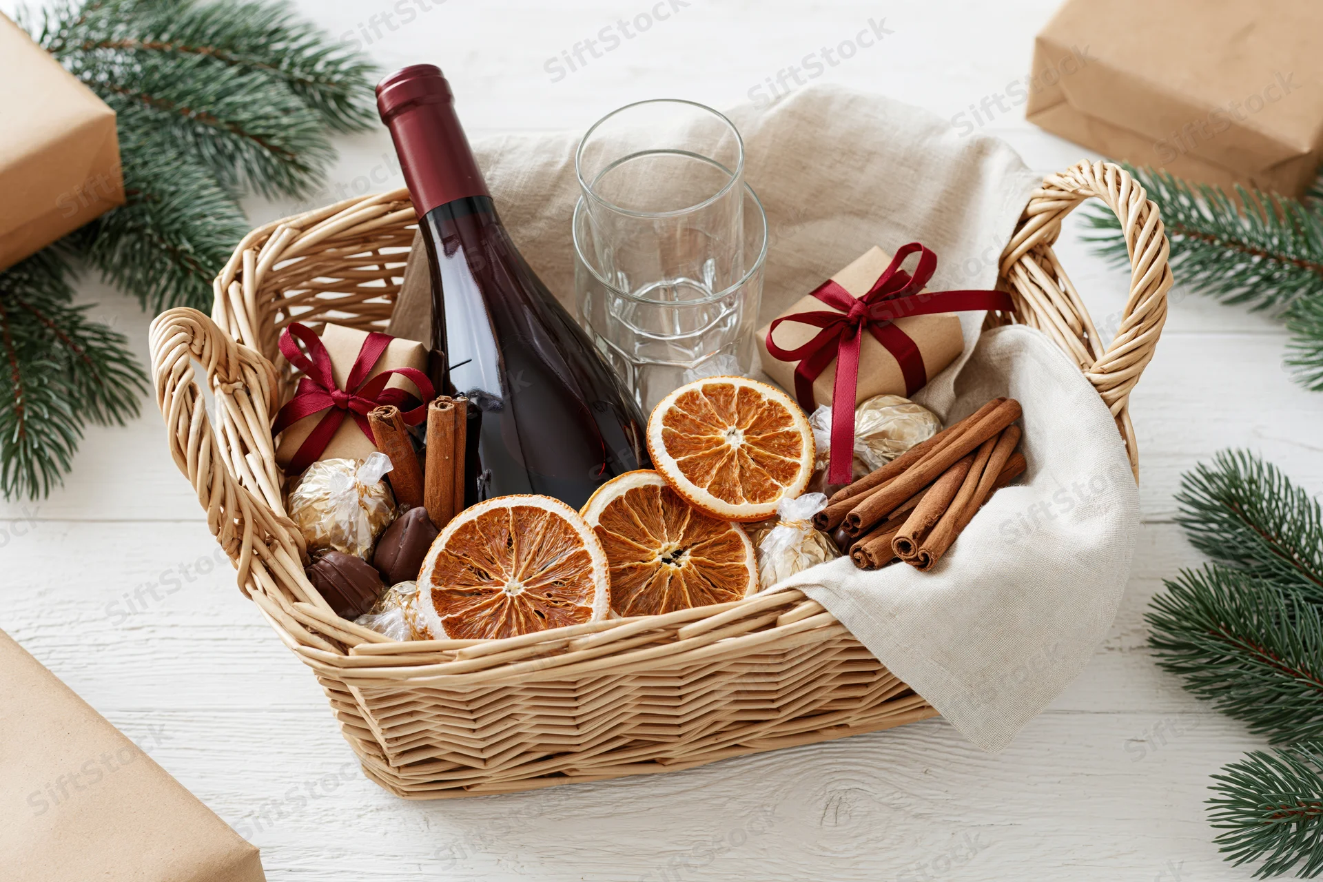 Wicker gift basket filled with red wine bottle, dried oranges, cinnamon sticks, chocolates, and wine glasses on white wooden table surrounded by pine branches and wrapped gifts