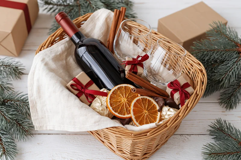 Wicker gift basket filled with red wine bottle, glasses, dried oranges, cinnamon sticks, and small wrapped gifts on white wooden table surrounded by pine branches