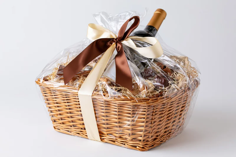 Wicker gift basket wrapped in cellophane with ribbon bow containing wine bottle and chocolates on white background