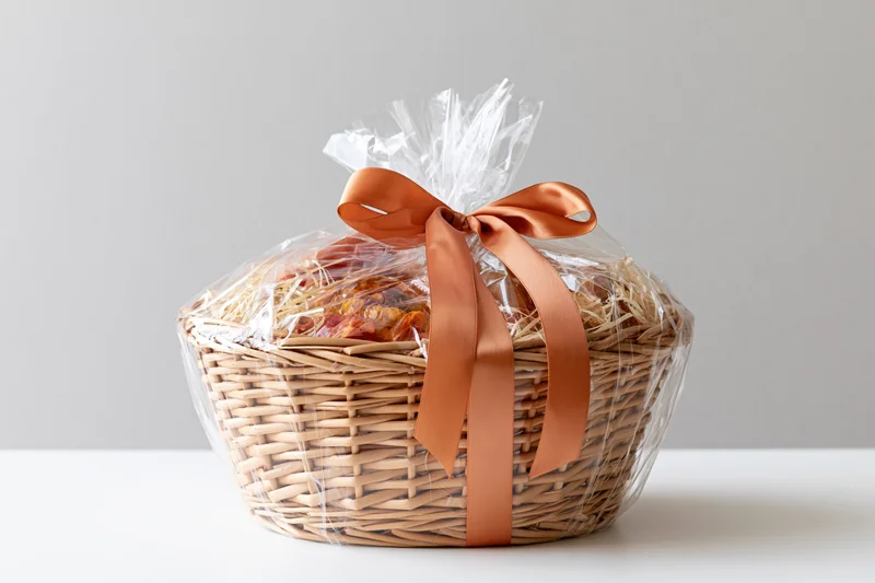 Wicker gift basket wrapped in cellophane with orange ribbon on white surface