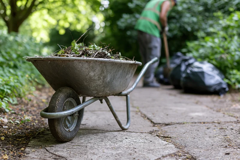 Metal wheelbarrow filled with garden debris on a cracked concrete path with a worker in the background using a shovel near black trash bags