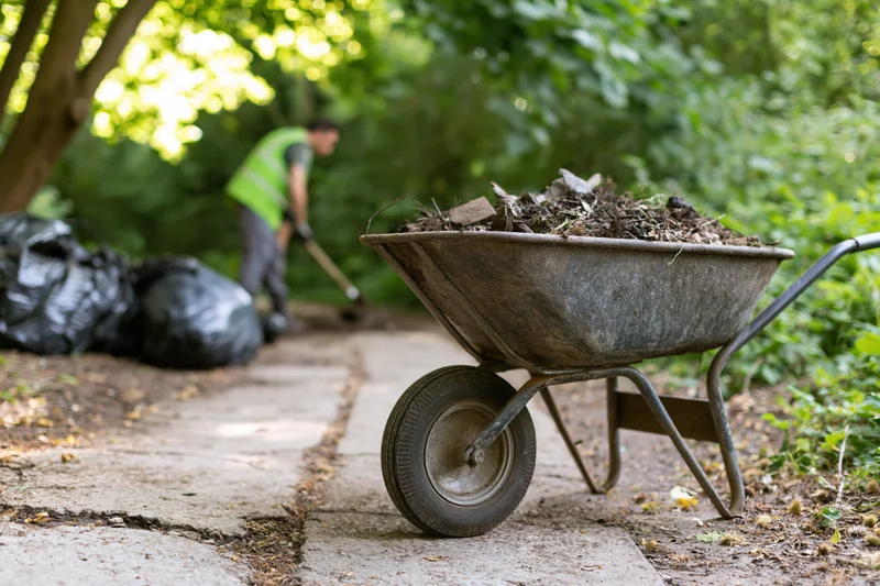 Metal wheelbarrow filled with garden debris on a cracked concrete path with a worker raking in the background