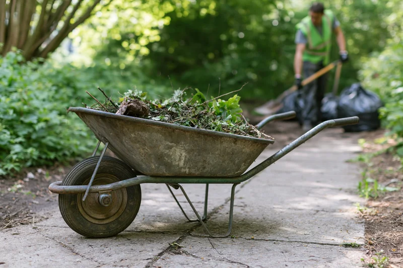 Metal wheelbarrow filled with garden debris on a cracked concrete path with a worker in the background using a shovel near trash bags