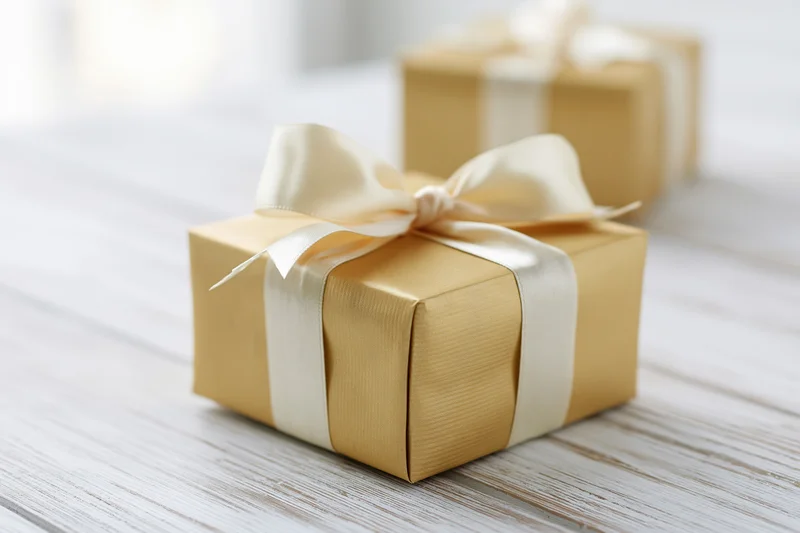 Gold-wrapped gift box with cream ribbon bow on white wooden table