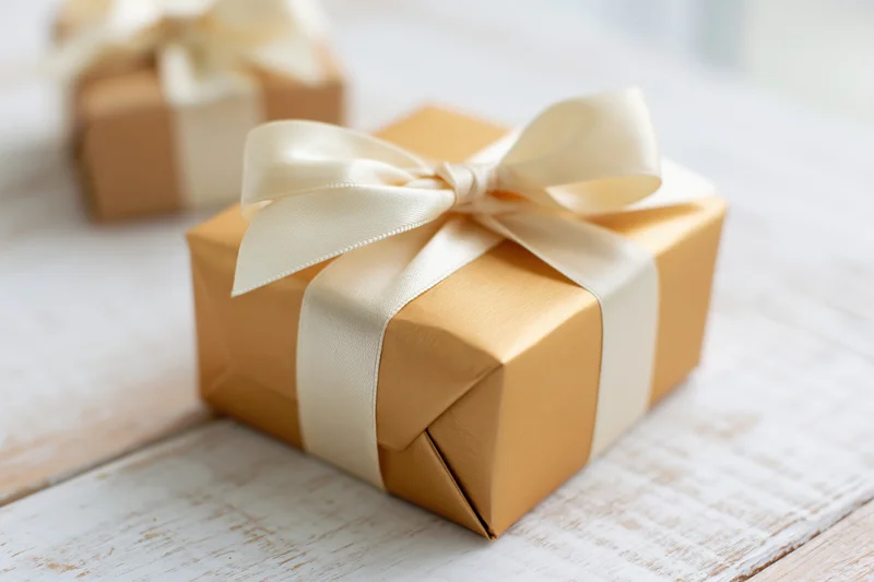 Gold-wrapped gift box with cream ribbon bow on white wooden surface