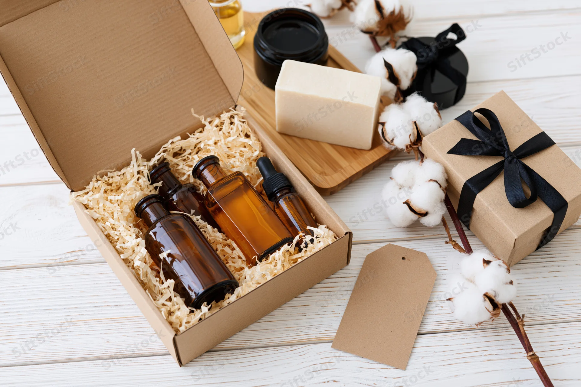 Open kraft gift box with amber glass bottles and natural soap on wooden surface surrounded by cotton plants and gift packaging