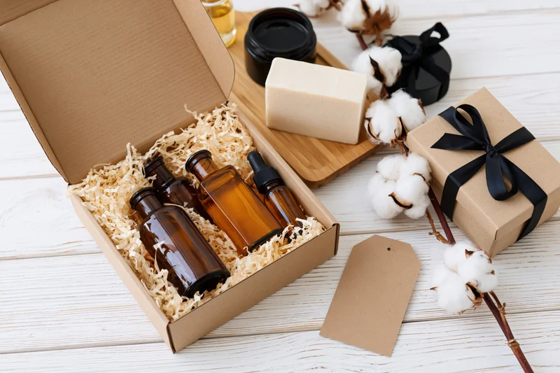 Open kraft gift box with amber glass bottles and natural soap on wooden surface surrounded by cotton plants and gift packaging