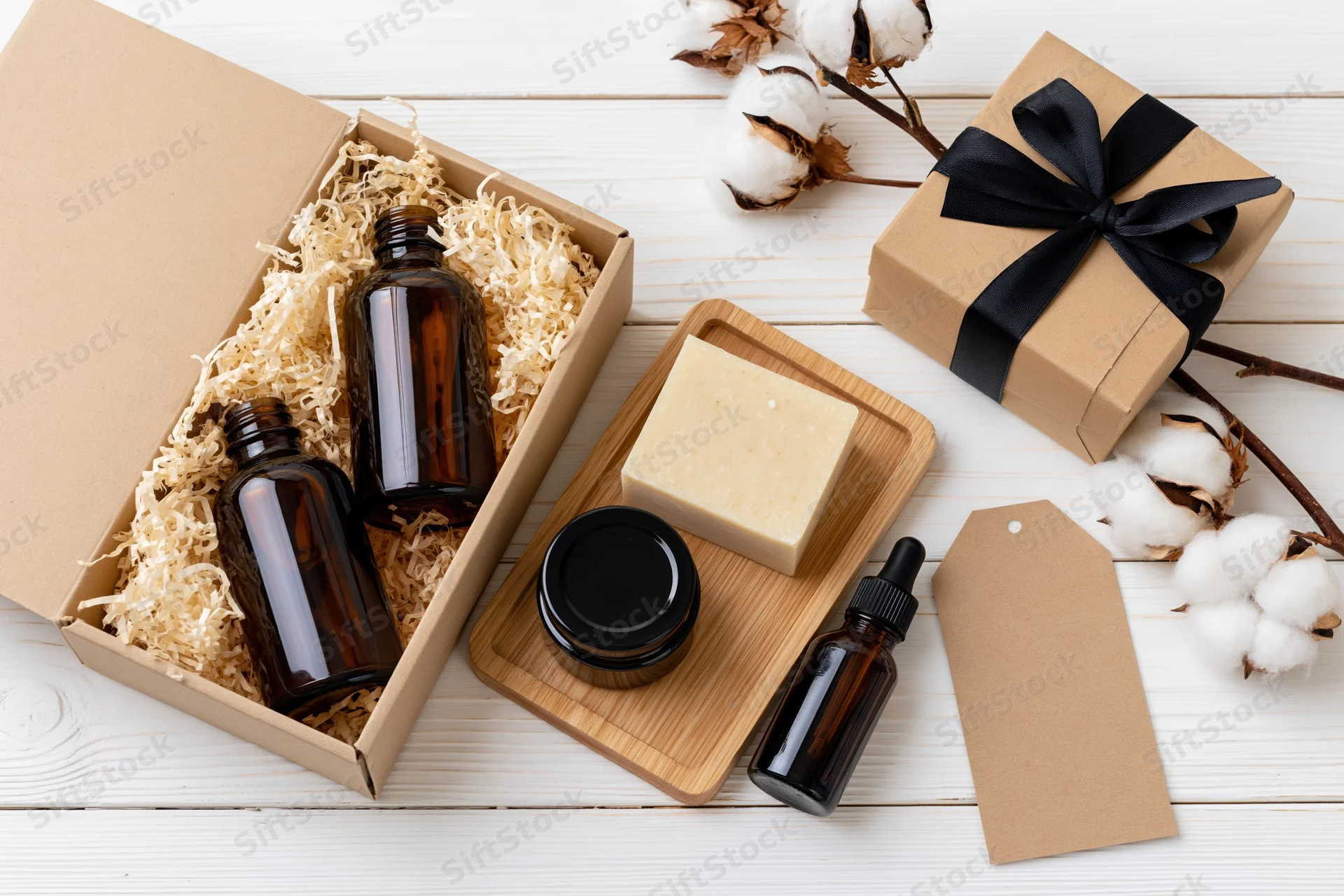 Open kraft gift box with amber bottles and soap on wooden tray beside cotton plants and wrapped gift