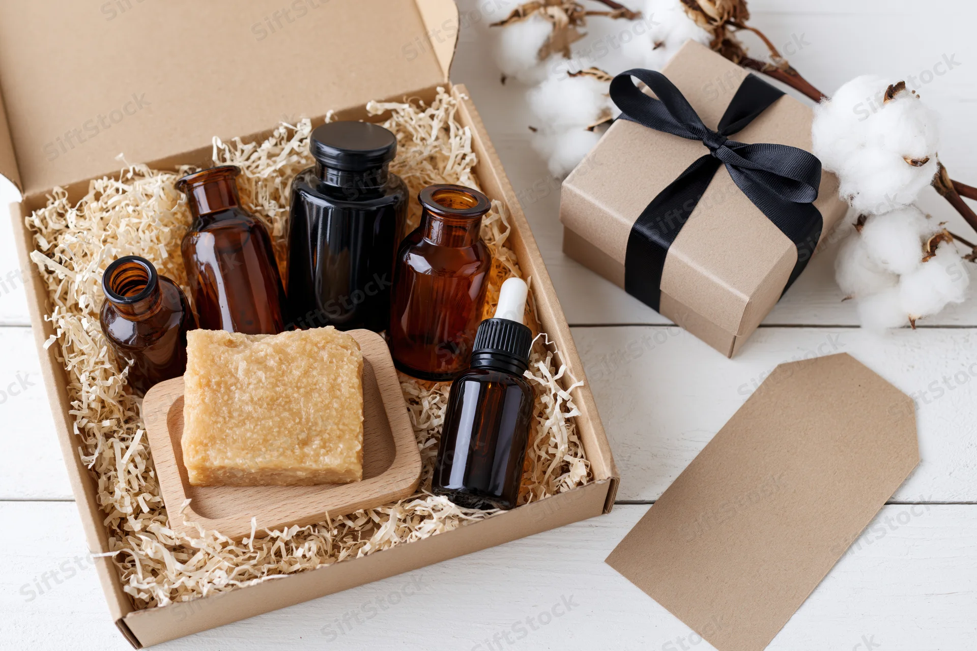 Open gift box with amber bottles and soap on shredded paper beside cotton plants and a wrapped gift