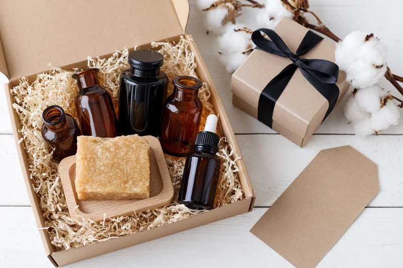 Open gift box with amber bottles and soap on shredded paper beside cotton plants and a wrapped gift