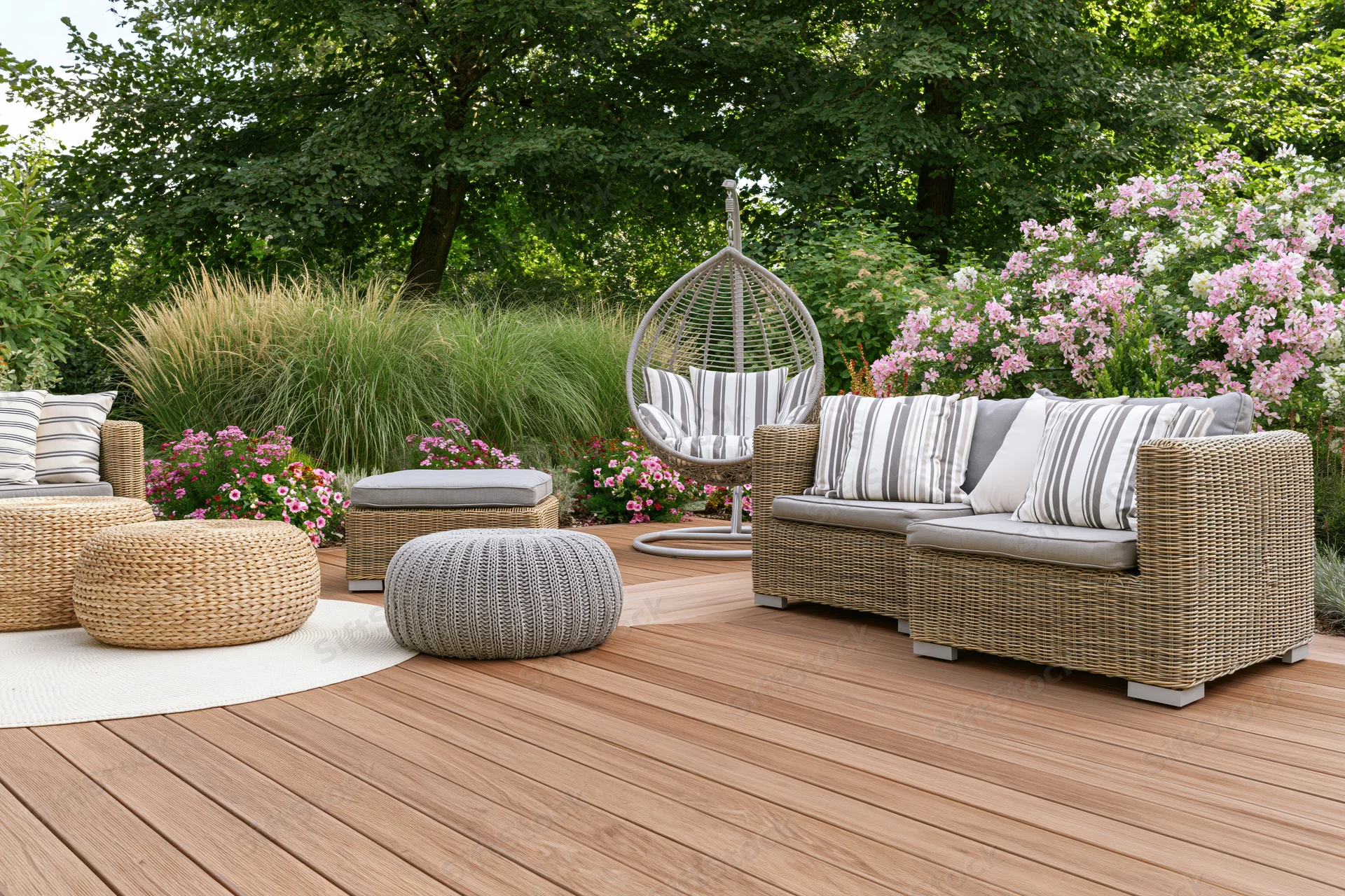 Rattan lounge furniture with striped cushions arranged on a wooden deck surrounded by flowering shrubs and greenery