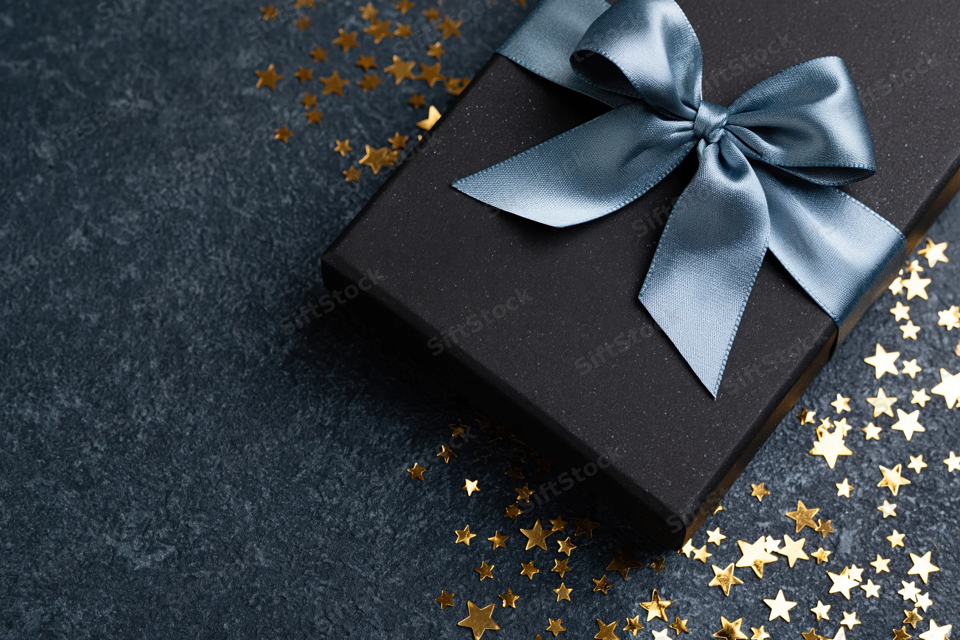 Black gift box with silver-blue satin ribbon bow on dark surface surrounded by gold star confetti