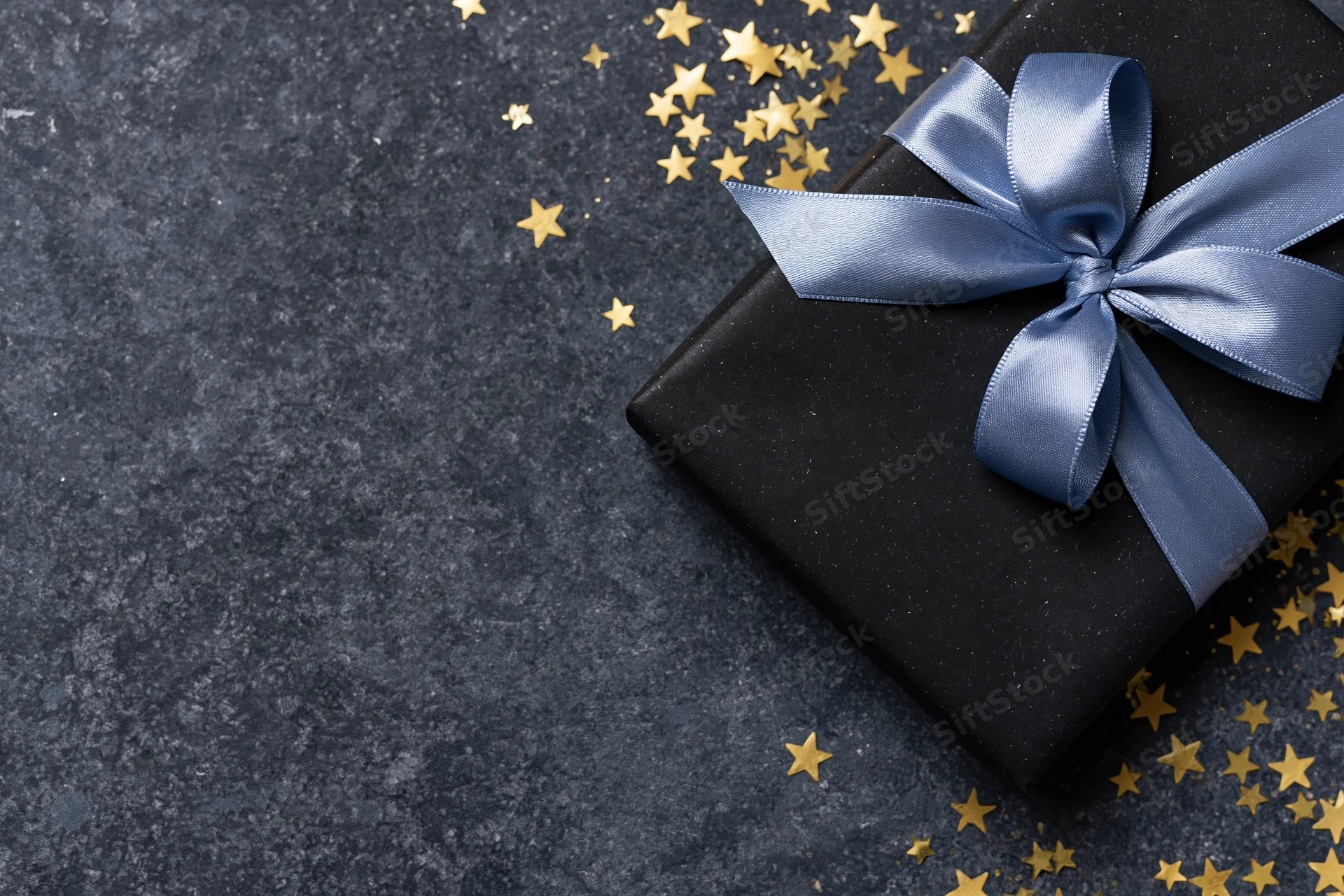 Black gift box wrapped with blue satin ribbon bow on dark textured surface with scattered gold stars and copyspace