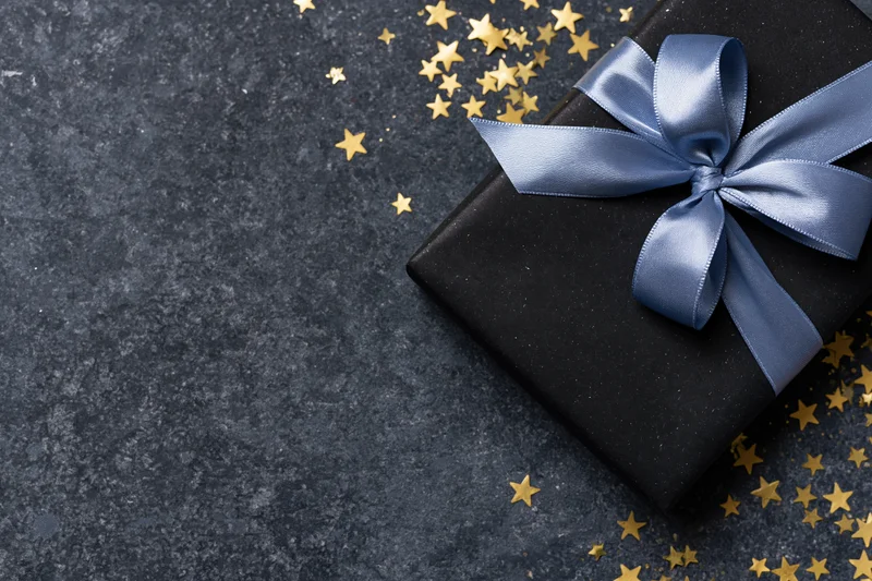 Black gift box wrapped with blue satin ribbon bow on dark textured surface with scattered gold stars and copyspace
