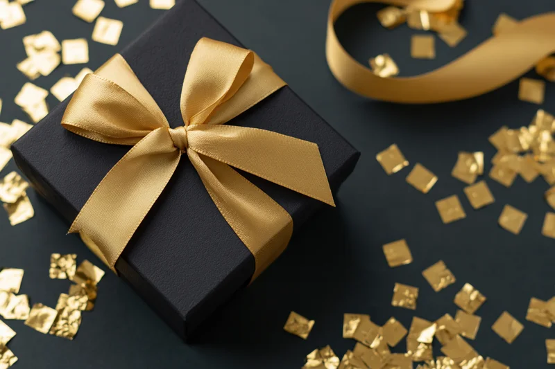 Black matte gift box with gold satin bow and scattered gold confetti on dark surface