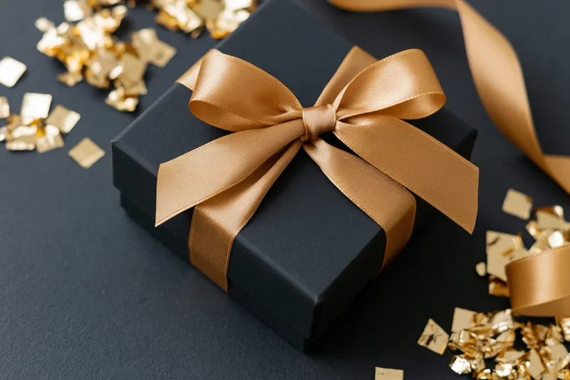 Black matte gift box with gold satin bow and scattered confetti on dark surface
