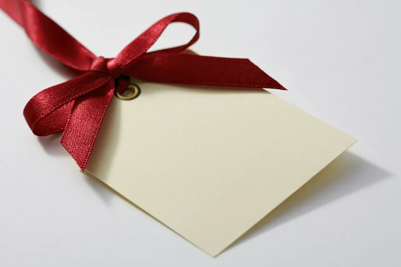 Cream gift tag with red ribbon bow attached by brass eyelet on white background