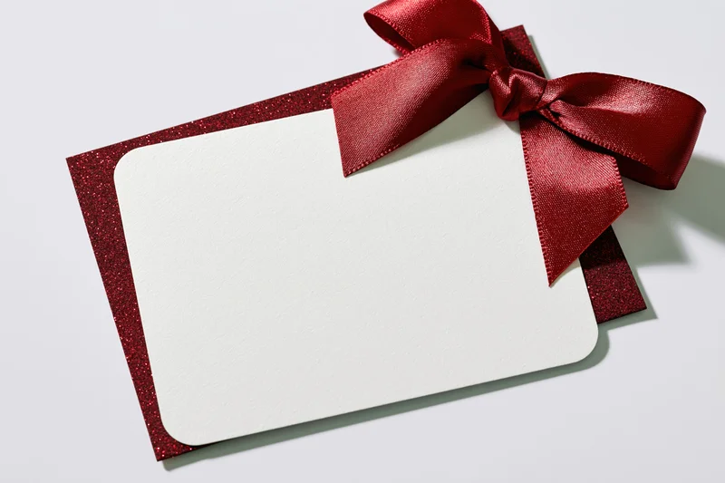 Blank white gift card with red glitter border and ribbon bow on white surface