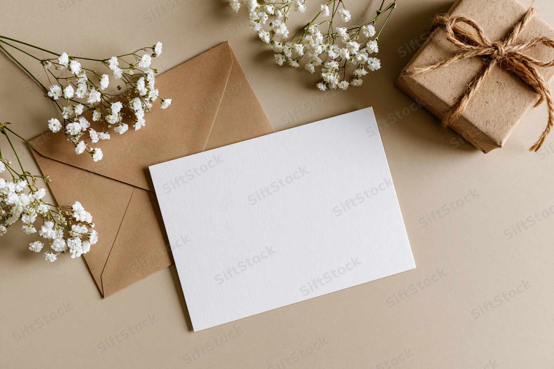 Blank white greeting card lying flat next to a kraft paper envelope and small gift box with baby's breath flowers on beige surface