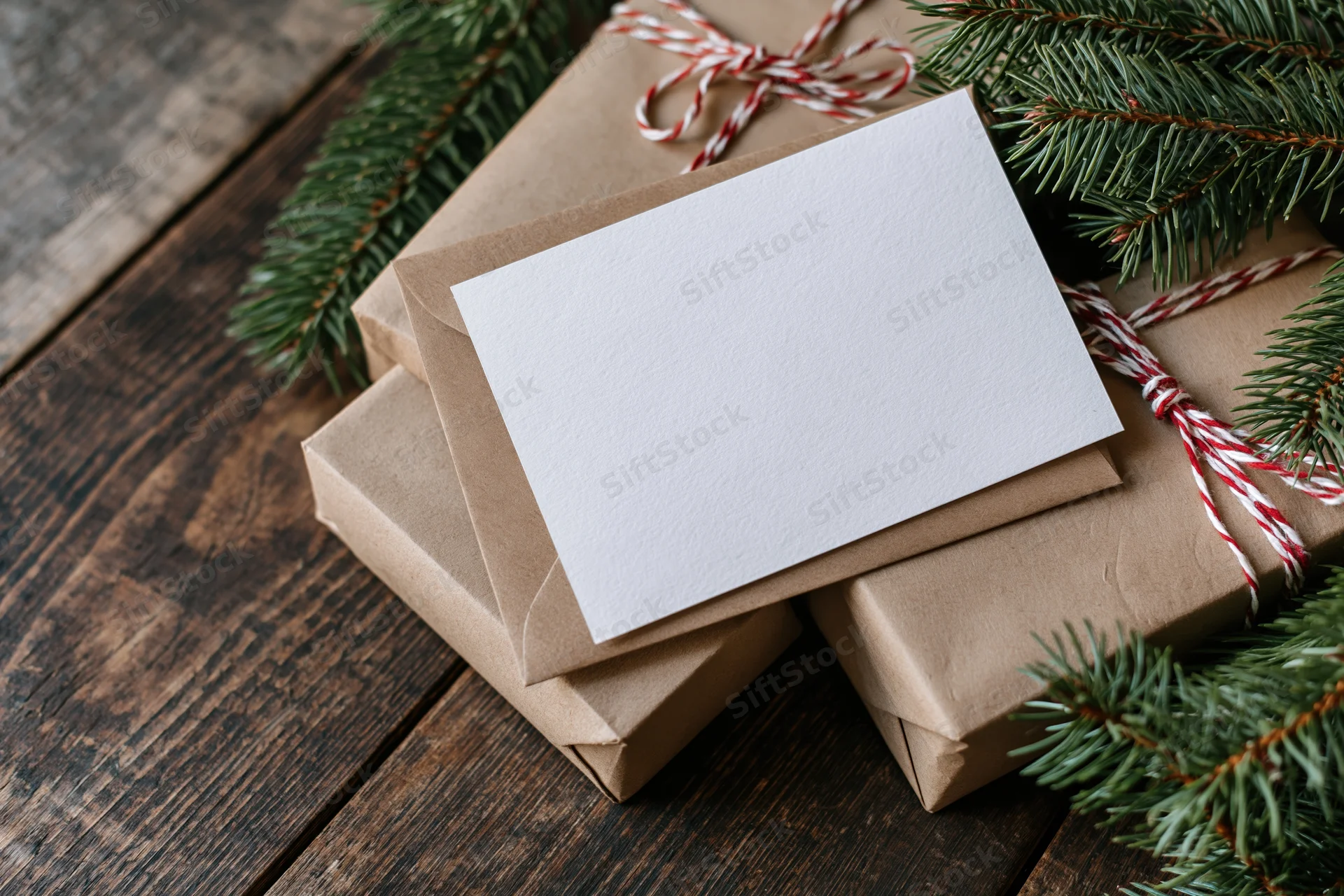 Blank white greeting card on kraft envelope with wrapped gifts and pine branches on wooden table