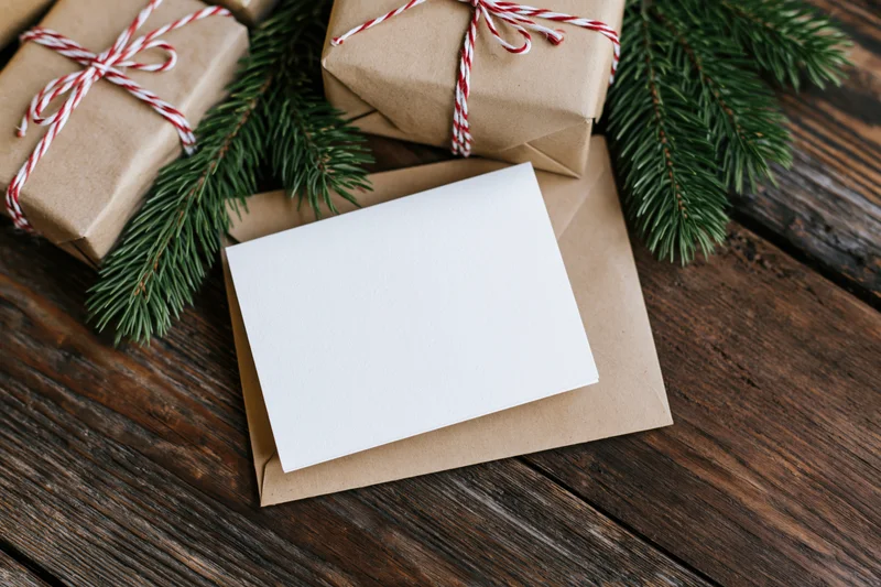 Blank white greeting card on kraft envelope with wrapped gifts and pine branches on wooden surface