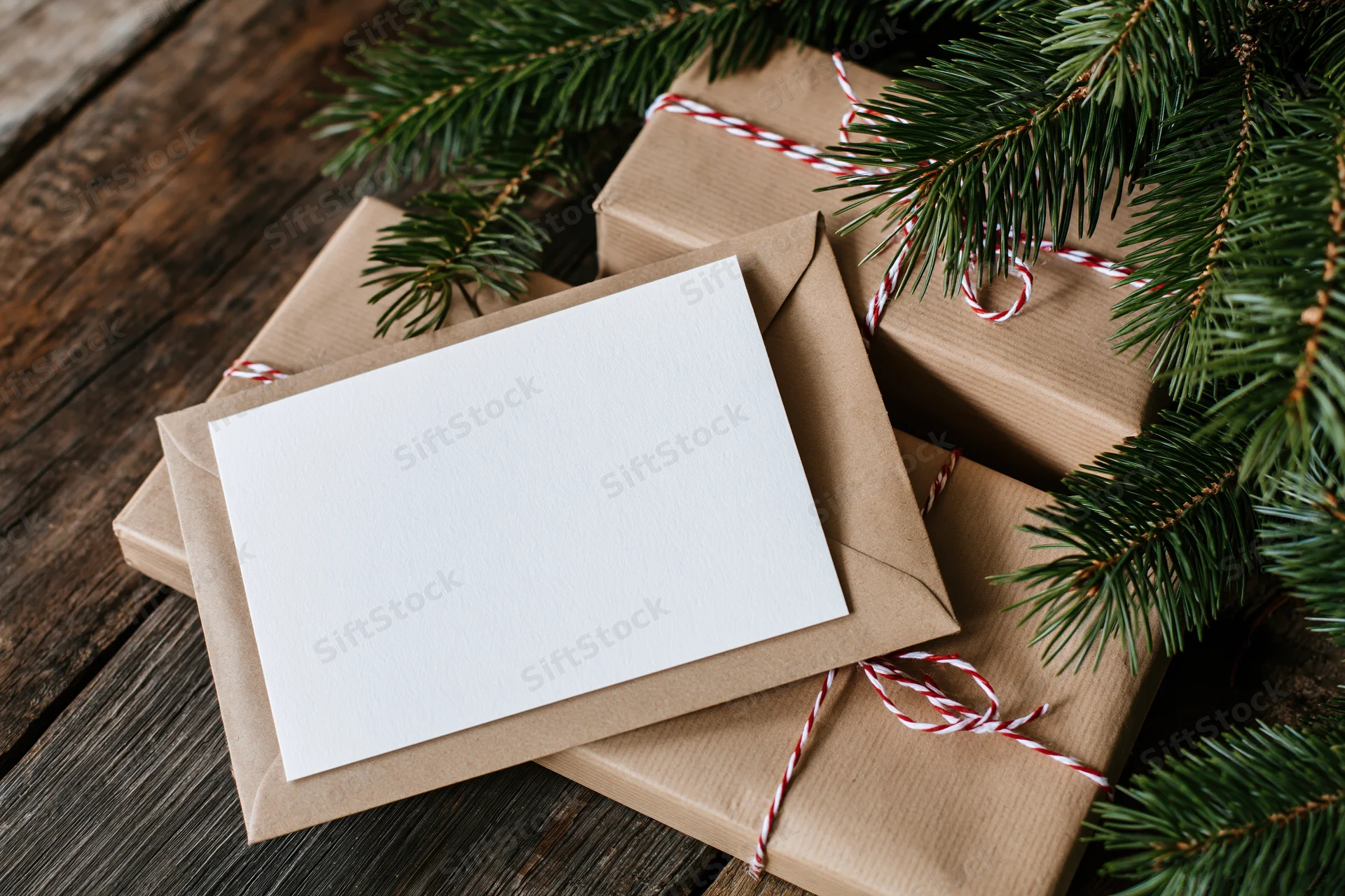 Blank white greeting card on kraft envelope with wrapped gifts and pine branches on wooden surface