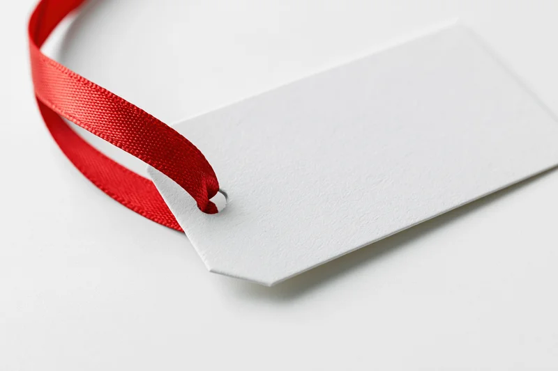 Blank white gift tag with red satin ribbon on white surface