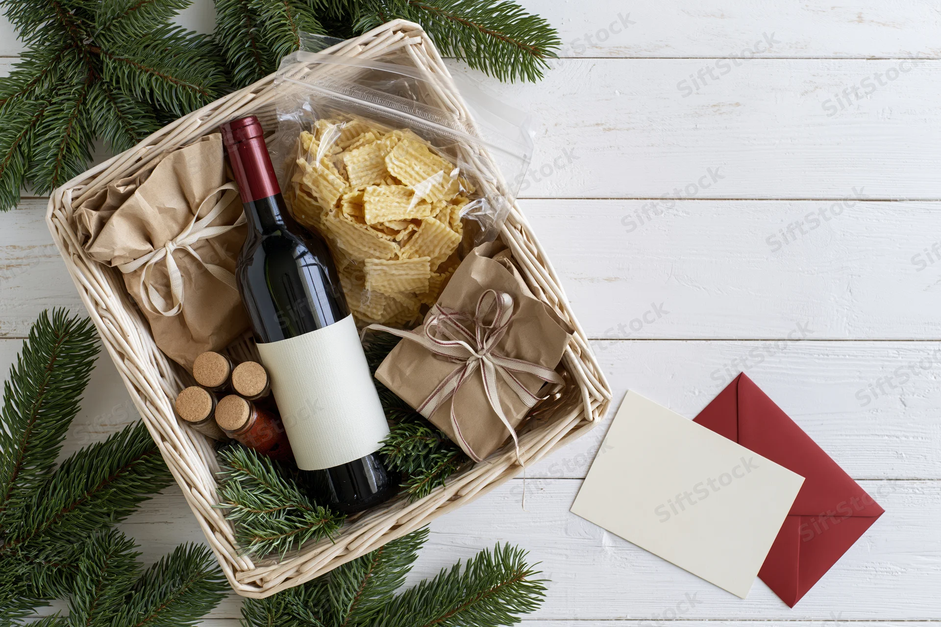 Christmas gift basket with wine and crackers on white wooden table, surrounded by pine branches and blank card with envelope