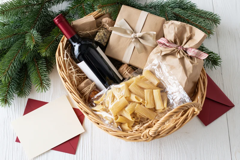 Christmas gift basket with wine and pasta on white wooden table surrounded by pine branches and envelopes