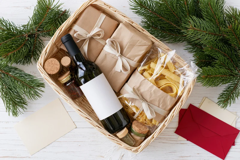 Christmas gift basket with wine, pasta, jars and wrapped gifts on white wooden table surrounded by pine branches