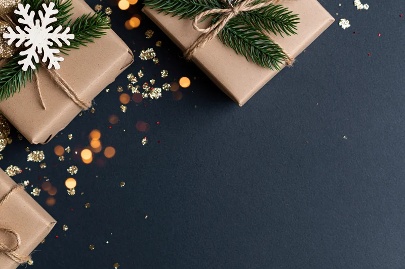 Christmas gift boxes wrapped in brown kraft paper with pine sprigs and snowflake ornaments on dark surface with bokeh lights and glitter scattered around