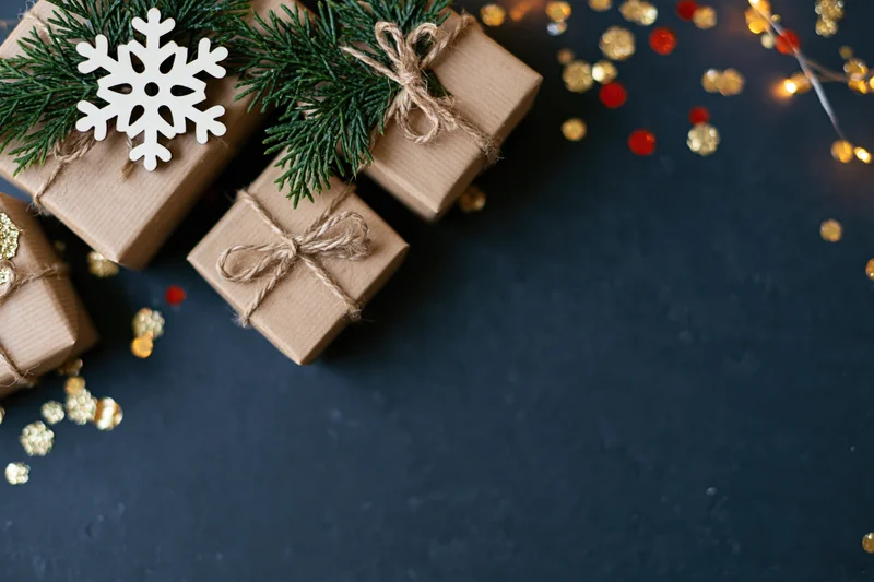 Christmas gifts wrapped in kraft paper with twine and pine sprigs on dark surface with string lights and confetti