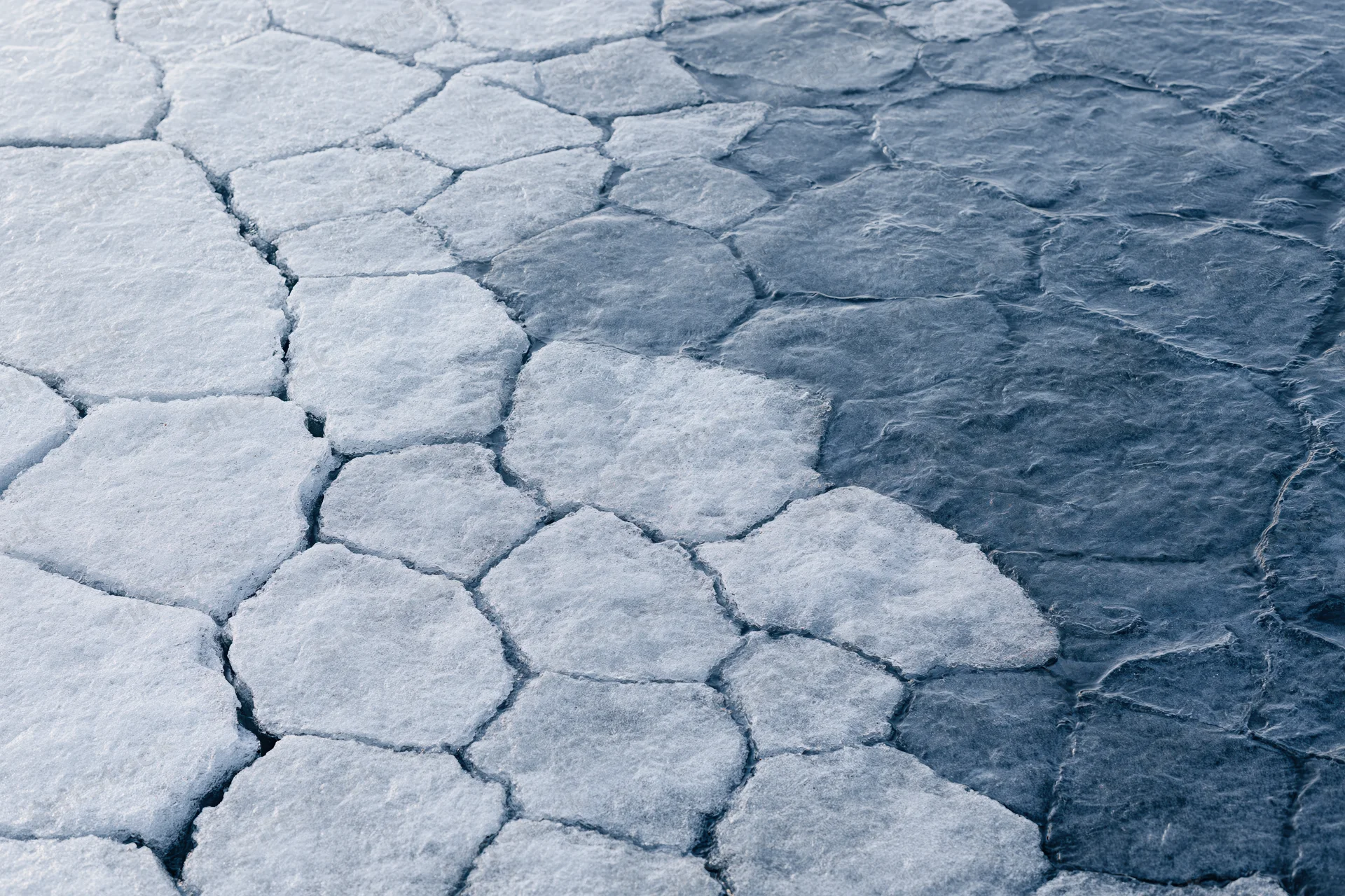 Fractured ice surface with hexagonal cracks and dark water patches, aerial view