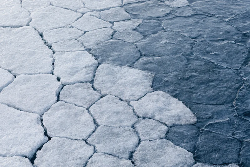 Fractured ice surface with hexagonal cracks and dark water patches, aerial view