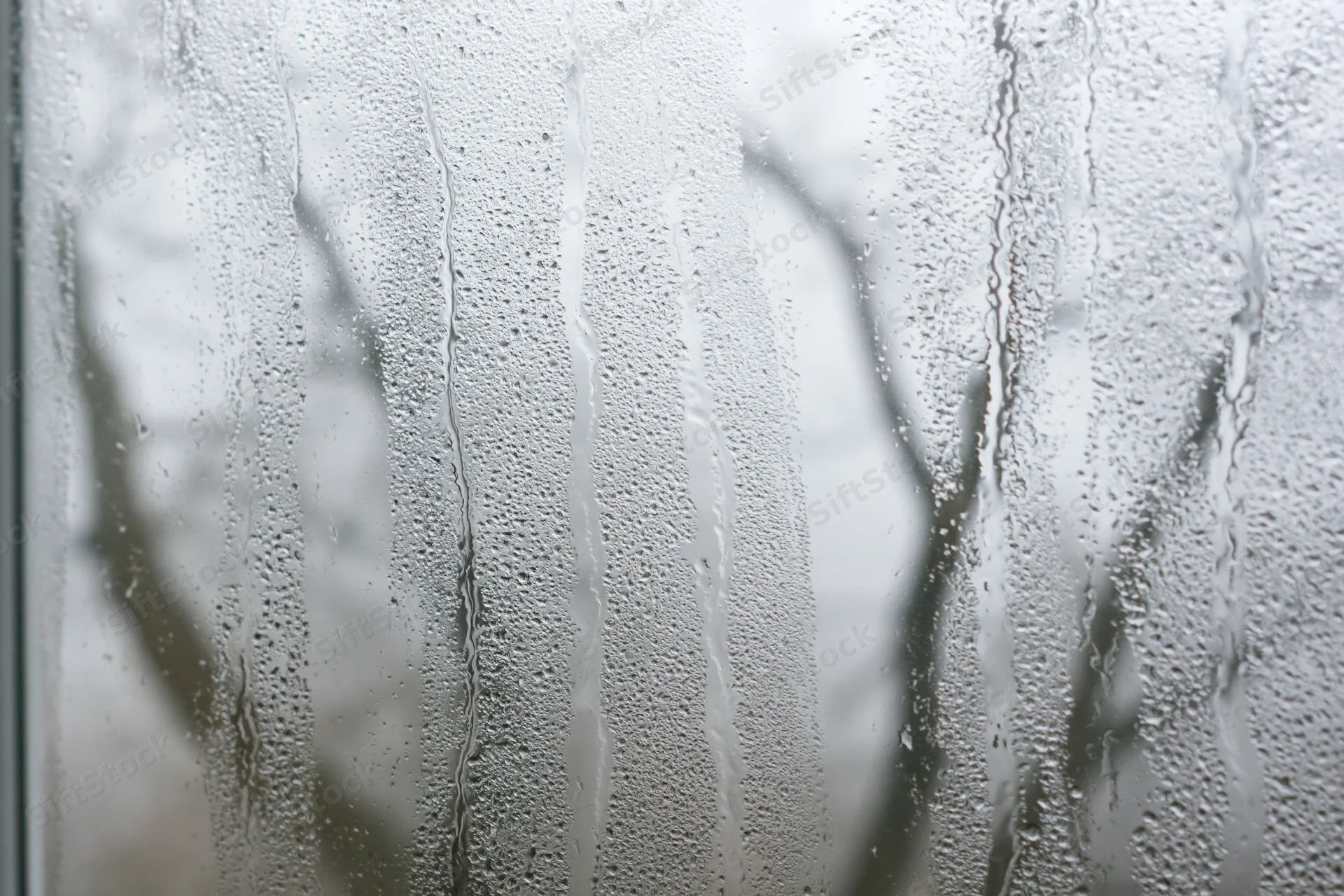 Close-up of a fogged window pane covered in heavy condensation with water droplets and streaks, showing blurred tree branches outside