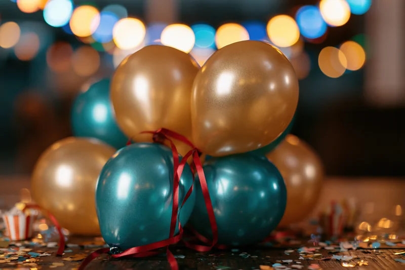 Cluster of metallic gold and teal party balloons with red ribbons on a wooden table surrounded by confetti and blurred festive lights