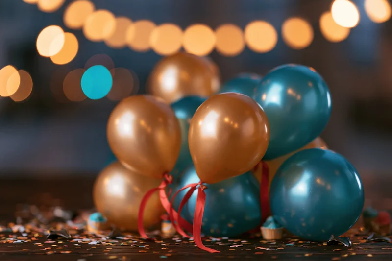 Metallic gold and teal balloons with red ribbons and confetti on a dark wooden table, string lights glowing in the background