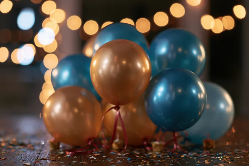 Cluster of metallic gold and teal balloons with red ribbons on a wooden floor scattered with confetti, illuminated by warm bokeh lights in the background