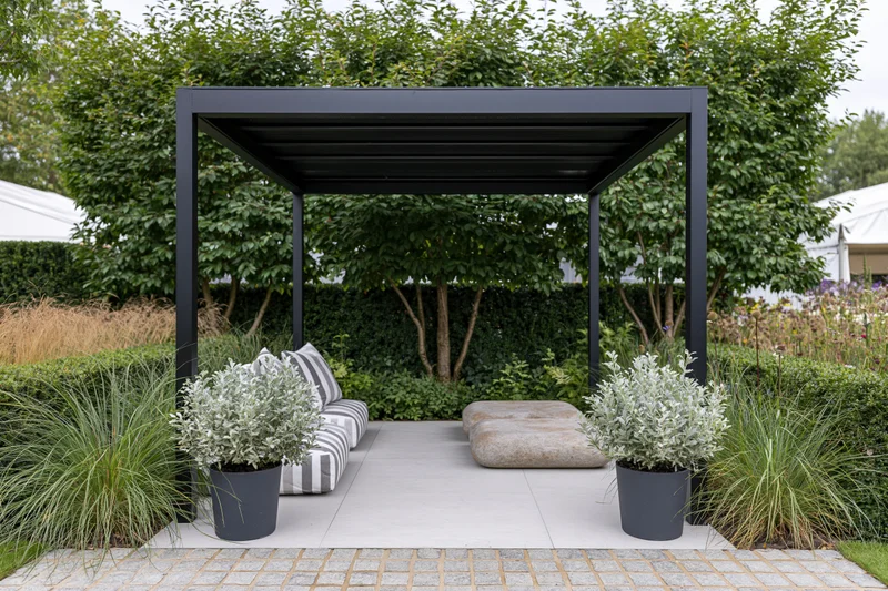 Black steel pergola over paved patio with striped cushions and potted plants in a landscaped garden