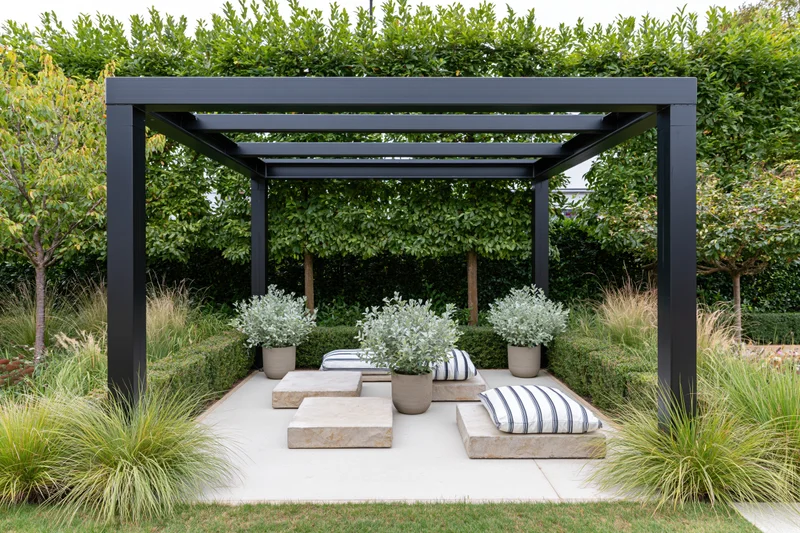 Black steel pergola over low stone seating with potted plants and striped cushions in a contemporary garden