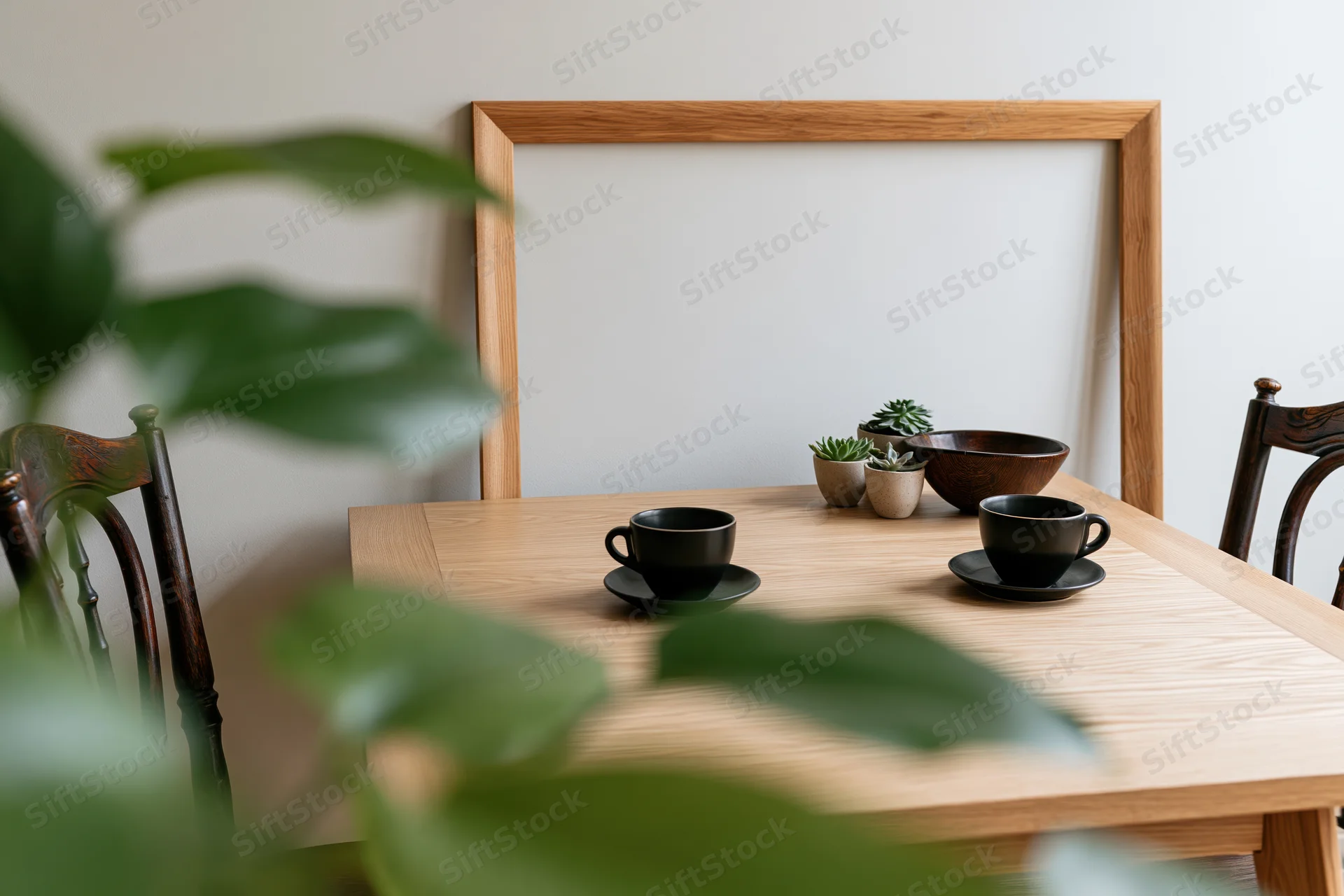 Wooden dining table set with black cups and saucers, small potted succulents, and a blank wooden frame leaning against the wall, viewed through blurred green leaves in the foreground