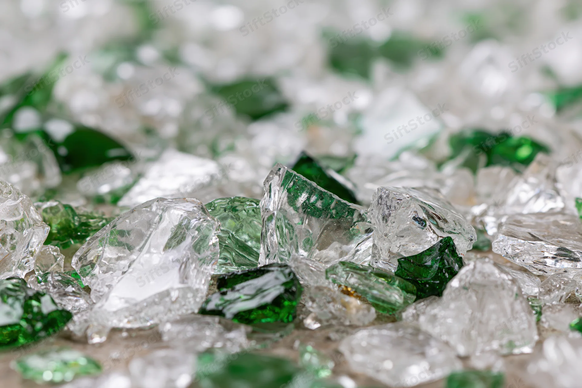 Broken glass shards scattered densely on a surface, some green and transparent, with sharp edges and reflective surfaces