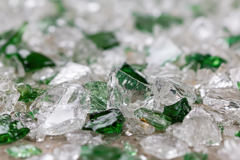 Broken glass shards scattered densely on a surface, some green and transparent, with sharp edges and reflective surfaces