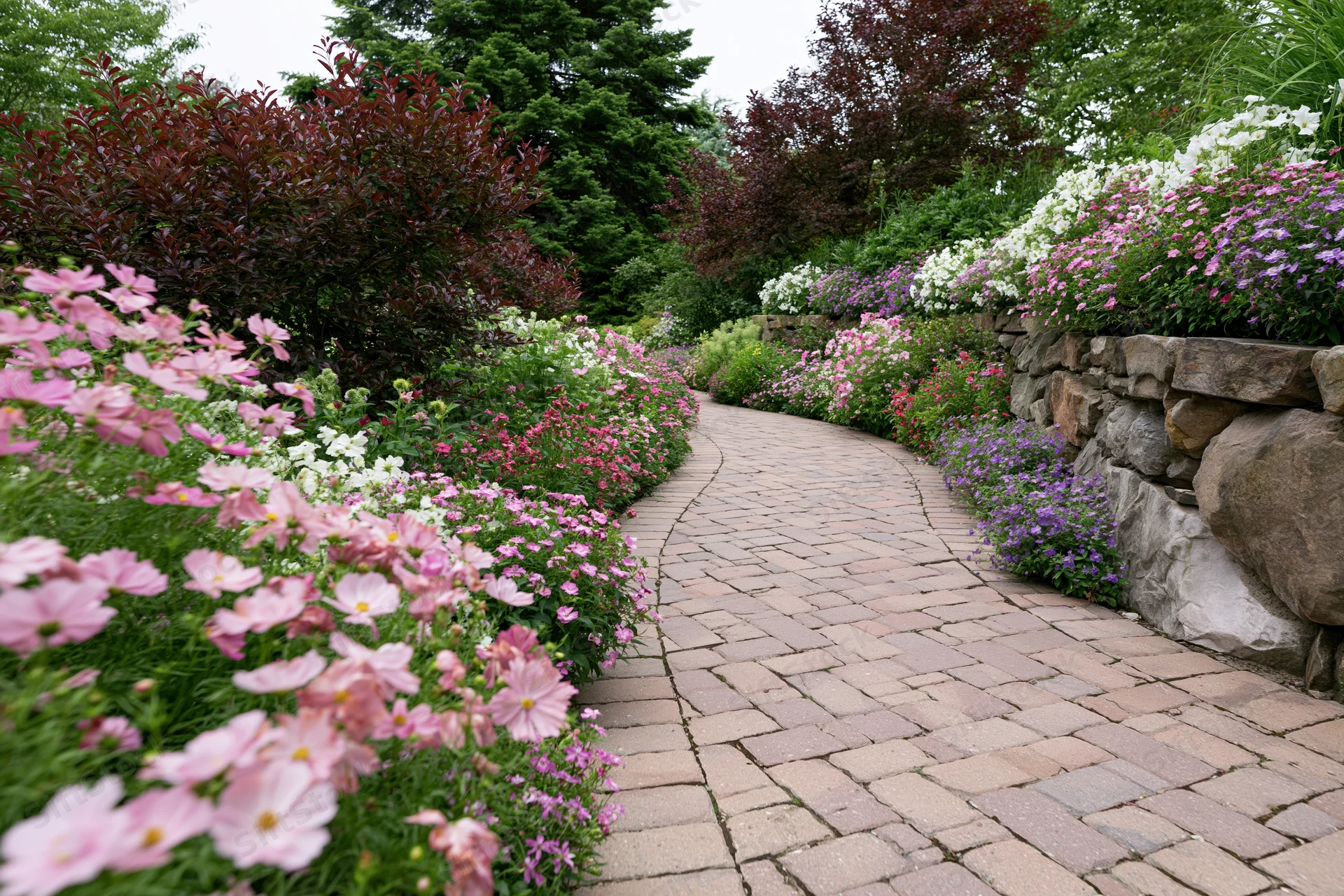 Curved brick paver path winding through flowerbeds bordered by stone wall and shrubs