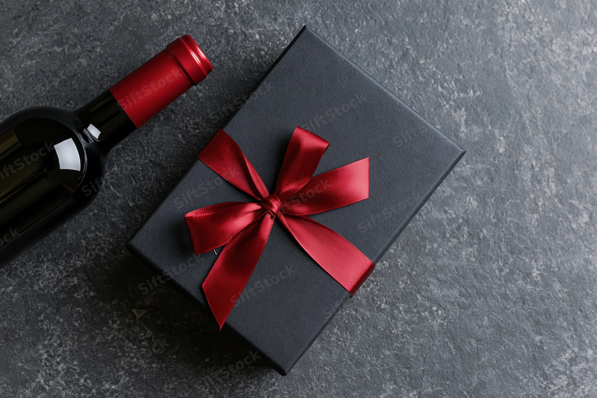 Dark gift box with red satin ribbon bow beside a wine bottle on stone surface